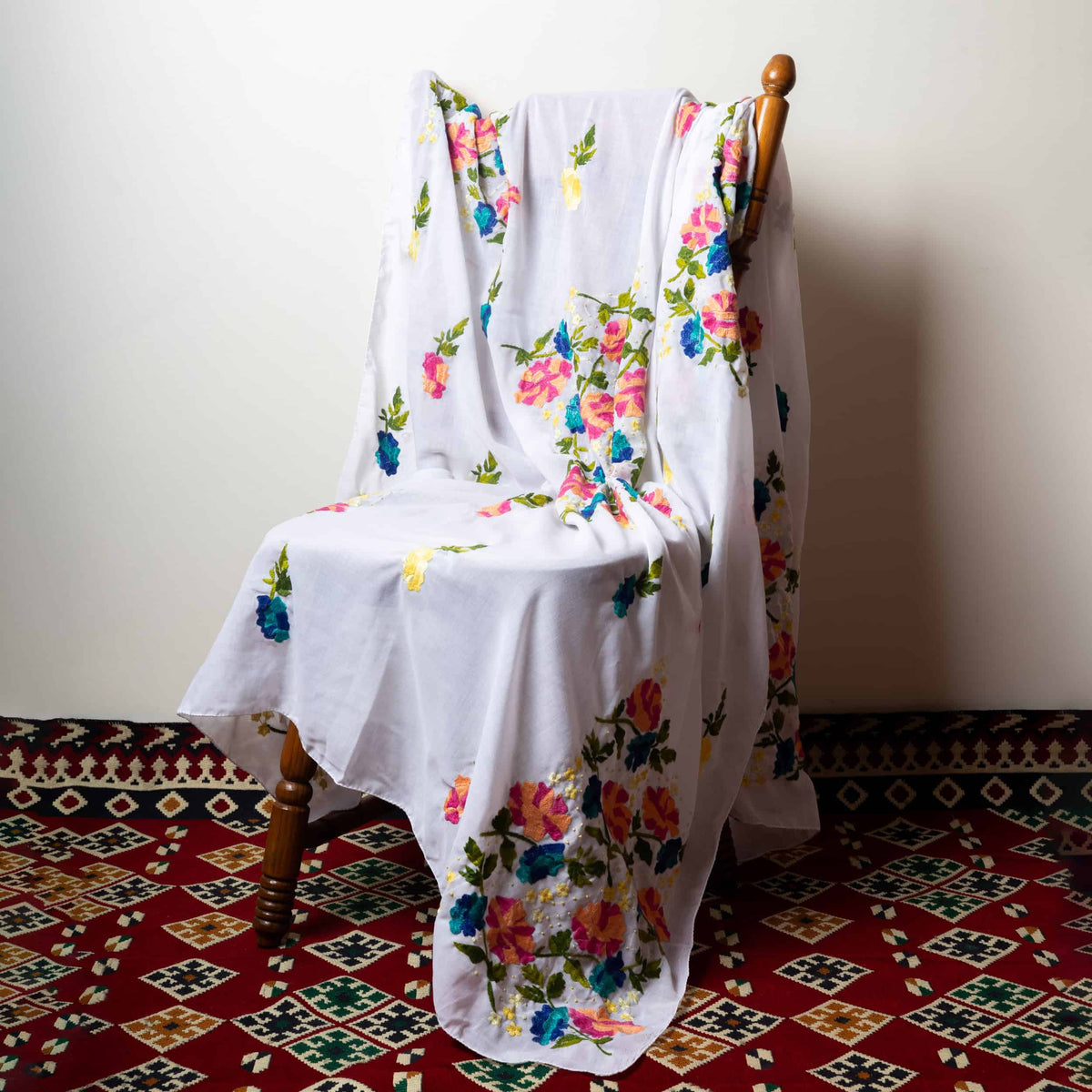 A white fabric with hand-embroidered floral patterns draped over a wooden chair against a floral background.
